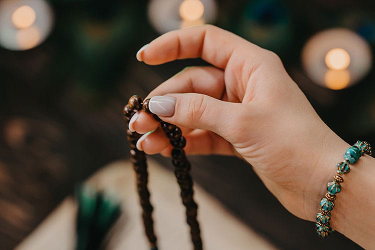 Close-up of a person holding traditional mala beads for mantra meditation, with candles and sacred text in the background.