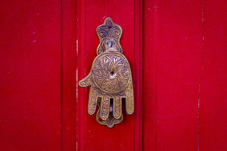 An ornate brass Hamsa-shaped door knocker mounted on a vibrant red wooden door. The design reflects traditional craftsmanship and symbolizes ancient protection across Jewish, Islamic, and Christian cultures.