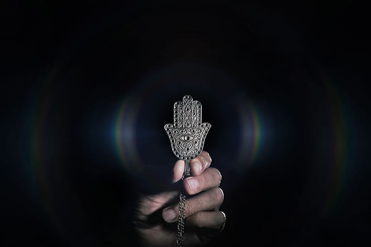 A hand holds a metallic Hamsa pendant against a dark background. The pendant features an eye in the center of the palm, symbolizing protection from the evil eye and negative energy.