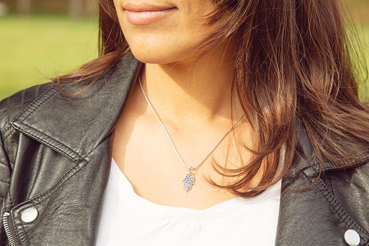A woman wearing a silver Hamsa pendant on a delicate chain, paired with a white shirt and black leather jacket. The scene captures a modern take on the ancient symbol, blending fashion with personal meaning.