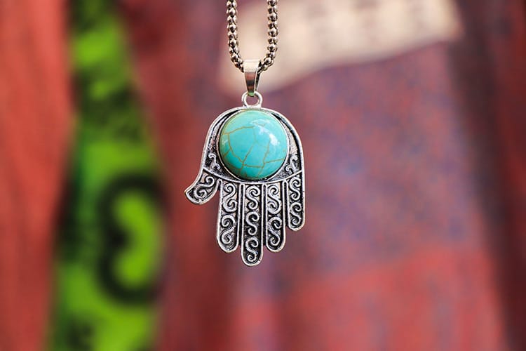 A close-up of a silver Hamsa pendant with a large turquoise stone in the center, hanging against a blurred background of patterned fabric. The image evokes personal meaning, protection, and spiritual grounding.