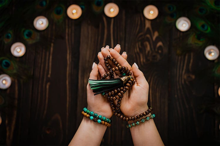 Hands gently holding wooden mala beads with green tassels, surrounded by gemstone bracelets and lit candles, symbolizing grounding, heart-centered intentions, and spiritual focus.