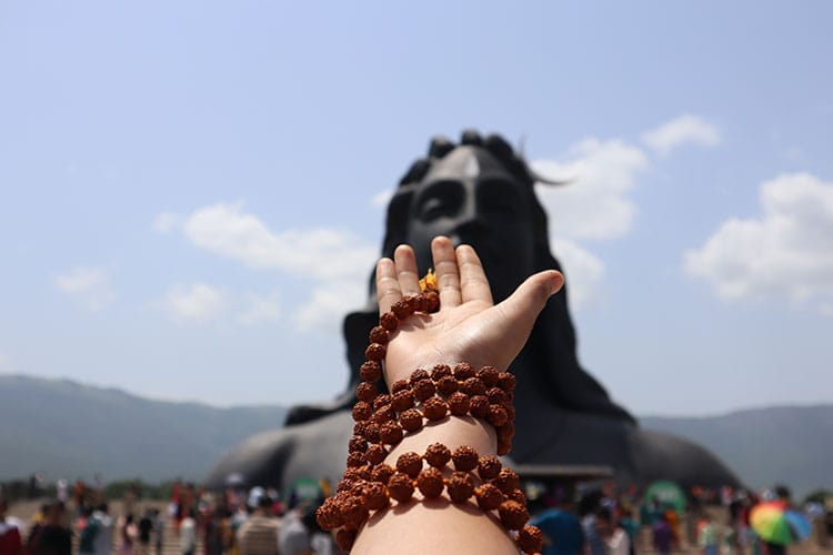 Hand adorned with Rudraksha malas extended toward a large statue of Lord Shiva. It symbolizes the sacred connection between Rudraksha beads and devotion in Hinduism.