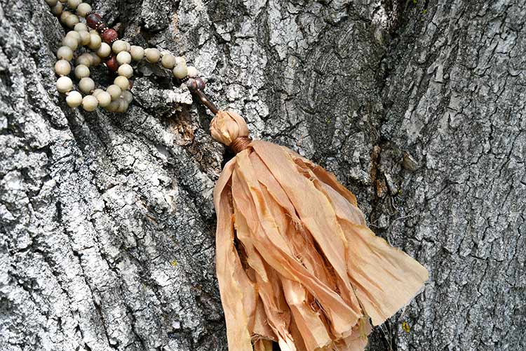 Mala beads with a large, rustic tan tassel resting against the textured bark of a tree, illustrating how tassels can add individuality and flow to a mala design.