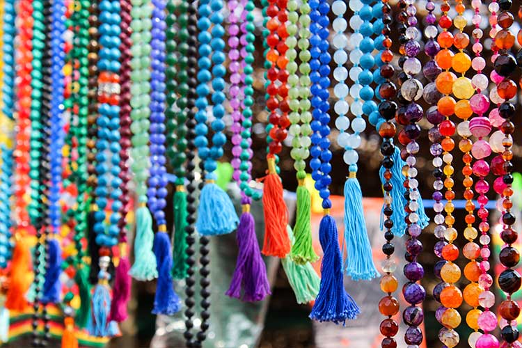 Colorful gemstone mala beads hanging in rows, featuring a variety of stones and vibrant tassels, symbolizing different energies like love, communication, and creativity.