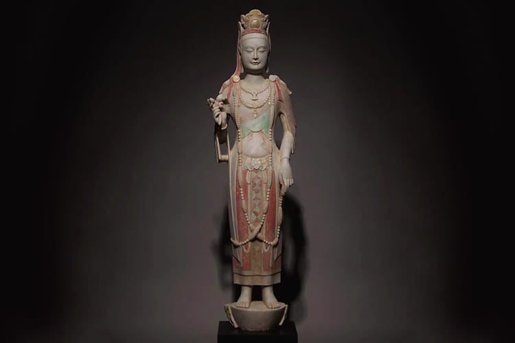 A tall, intricately carved Kuan Yin statue stands in soft spotlight. She wears flowing robes with subtle faded colors.