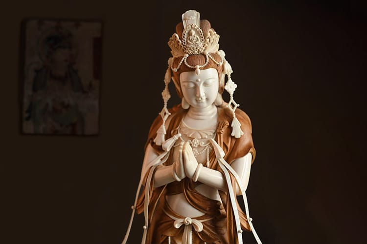 A finely detailed Kuan Yin statue made of white and warm-toned material. Her hands are in the namaste mudra gesture.
