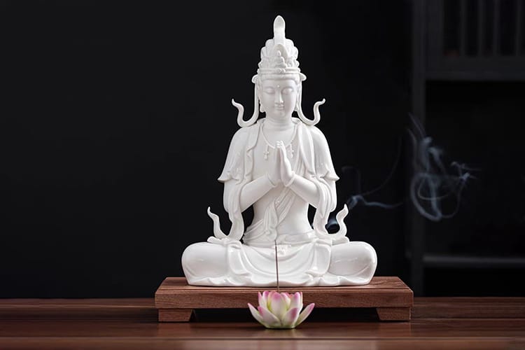 A serene white Kuan Yin statue sits in a meditative pose on a wooden base, hands in prayer, with incense smoke gently curling beside her.