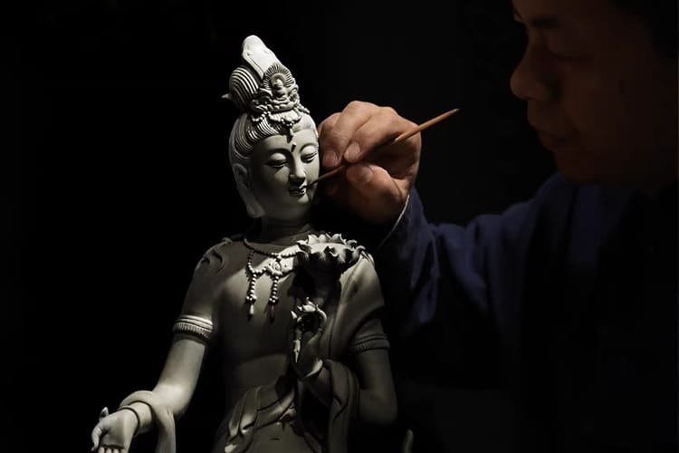 An artisan carefully paints the delicate face of a Kuan Yin statue. It highlights the craftsmanship and spiritual intention behind the sculpture.