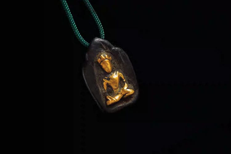 A dark pendant featuring a seated Buddha figure inlaid with gold. It is suspended on a green cord—an example of Indian Buddhist jewelry from the Kushan or Gupta era.