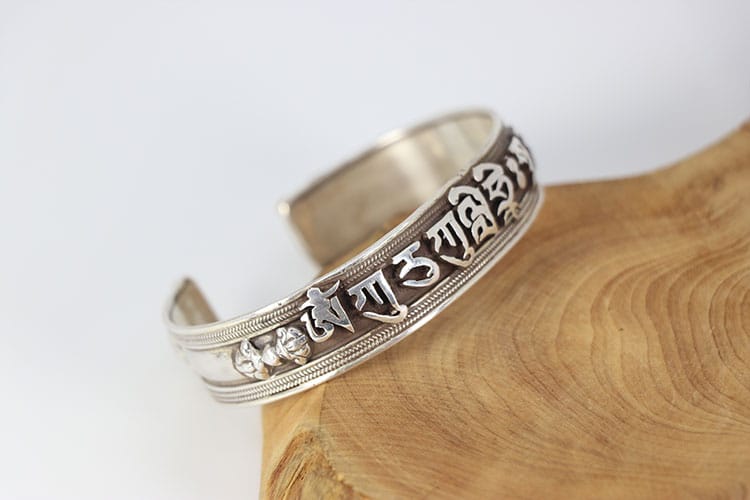 A silver Nepalese bracelet with darkened script and detailed carvings, resting on a wooden surface. It illustrates the use of blackened silver to create contrast, depth, and sacred meaning in Buddhist jewelry.