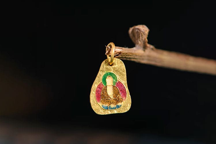 A small gold pendant depicting a seated Buddha in red, green, and blue lacquer. It hangs from a twig—exemplifying Burmese artistry through lacquer work.