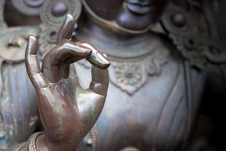 Close-up of a bronze Buddha statue with its hand forming a mudra. The background reveals intricate carvings on the statue's chest and robe.