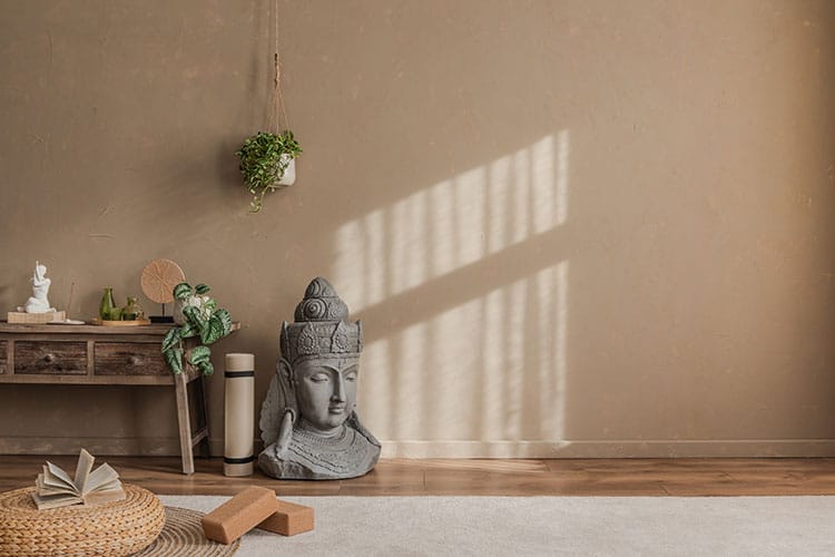 A peaceful meditation space with earthy tones and natural light filtering in through a window. A large sculpted Buddha head rests on the floor near a mat, yoga blocks, and an open journal.