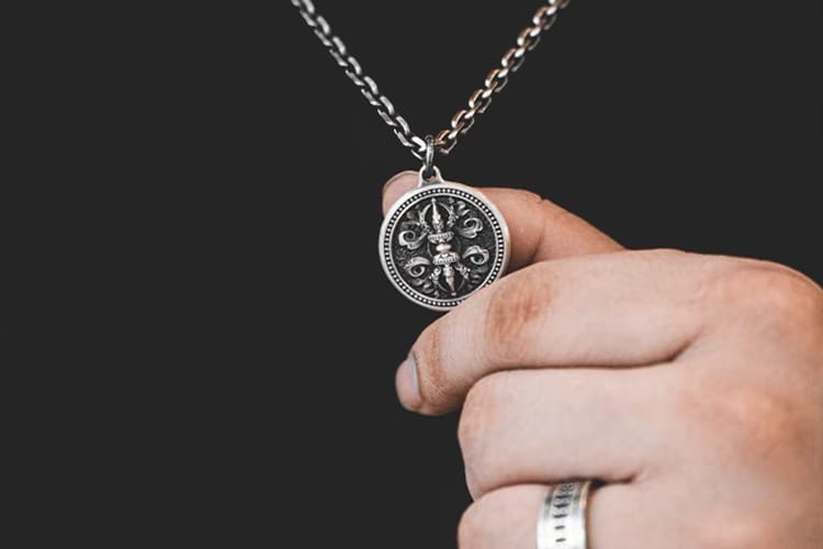 A close-up of a hand holding a silver Vajra pendant on a chain. The intricate design features symmetrical spokes around a central sphere, symbolizing unity, balance, and spiritual resilience.