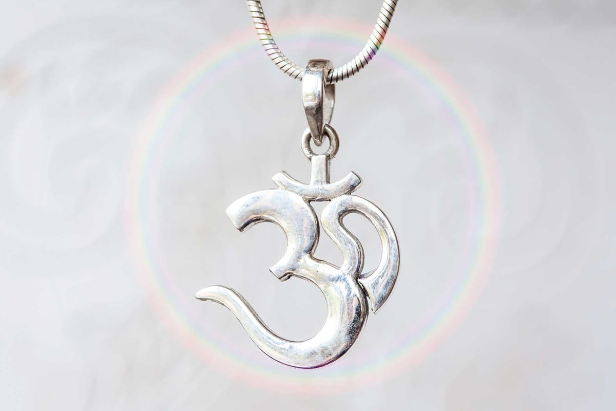 Close-up of a silver Om pendant hanging from a chain, with a soft rainbow ring in the background, symbolizing the sacred vibration of Aum and its connection to the infinite.