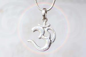 Close-up of a silver Om pendant hanging from a chain, with a soft rainbow ring in the background, symbolizing the sacred vibration of Aum and its connection to the infinite.