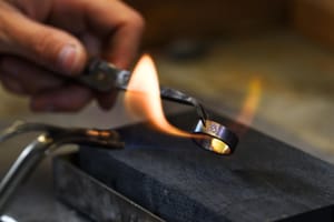 Close-up of an artisan using a flame to craft a metal ring engraved with the Om symbol. It captures the passion, tradition, and spirit behind Buddhist jewelry-making.